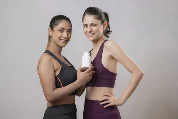 İki muhteşem dişi ayakta, gülümsüyor ve elinde vitamin şişesini sergiliyor.