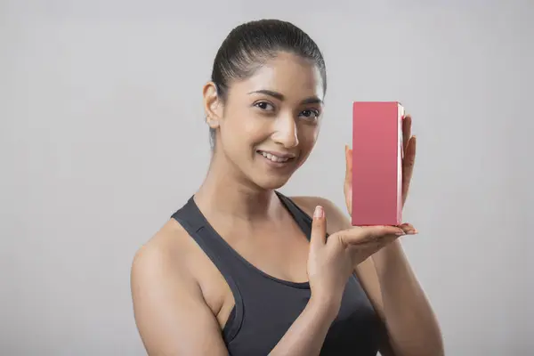 Güzel bir kadın gri bir eşofman giyiyor ve kameranın önünde kırmızı ilaç kutusu gösteriyor.