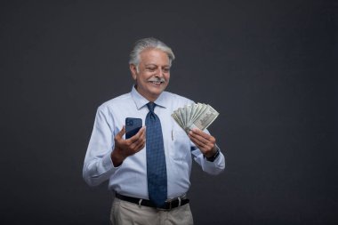 Yaşlı bir işadamı gülümsüyor, başka bir elde telefon tutarken parasına bakıyor.