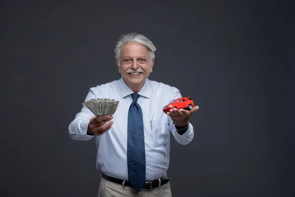 Ellerinde oyuncak araba ve para tutan yaşlı Hintli beyefendi ve kollarını açar ve parayı ve oyuncak arabayı başkasına verir.