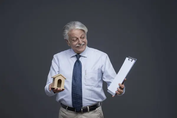 Yaşlı bir adam bir elinde ev oyuncağı tutarken gülümsüyor ve panosuna bakıyor.