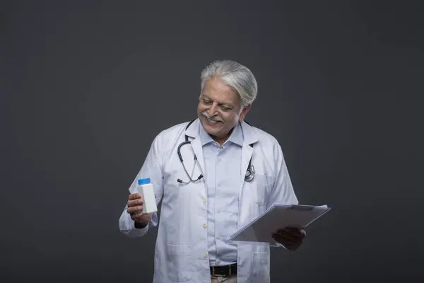 Paltolu ve steteskoplu bir doktor, bir elinde bir pano, diğer elinde ilaç şişesine bakıyor.