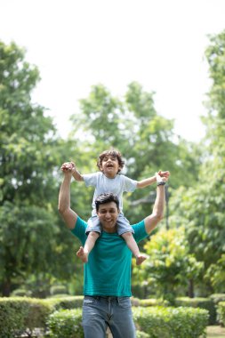 Hintli oğul dışarıda babasının omuzlarında otururken sırıtıyor.