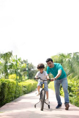 Meraklı çocuk parkta bisiklet sürmeyi babasından öğreniyor.
