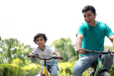 Baba ve oğul parkta bisiklet sürerken sırıtıyorlar.