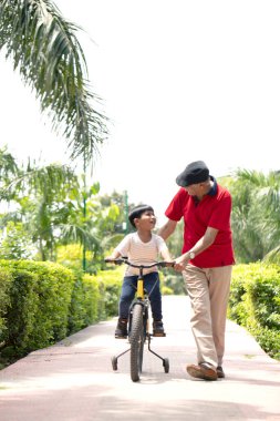 Adam torununa parkta bisiklet sürmeyi öğretiyor.