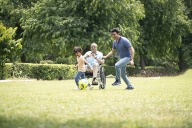 Oğlu ve torunu futbol oynarken izleyen yaşlı bir beyefendi.