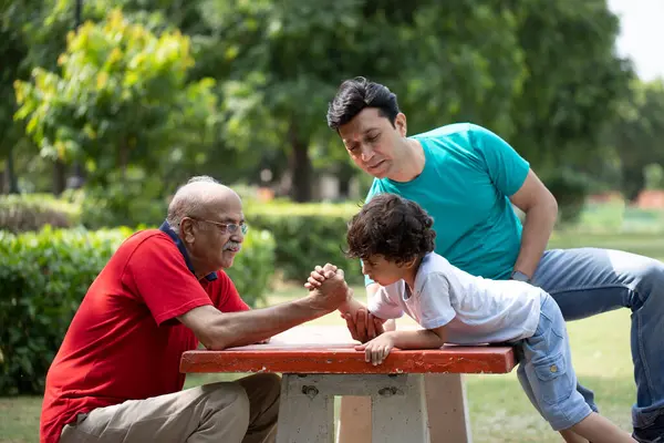Büyükbaba ve torun bilek güreşi yapıyor ve babam bahçede otururken onlara bakıyor.