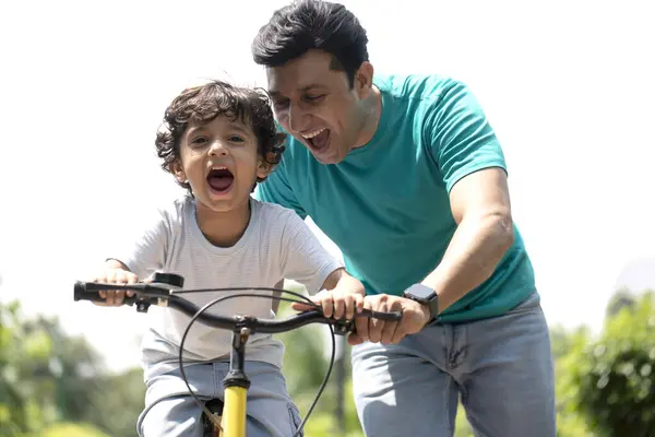 Parkta yakışıklı bir çocuk babasıyla bisiklet sürmeyi öğreniyor.