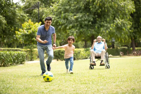 Yaşlı adam oğlunu ve torununu futbol oynarken izliyor.
