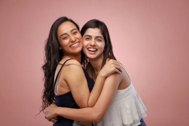 Güzel bayanlar pembe bir arka planda sarılıp sırıtıyorlar.