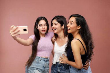 Üç sevimli arkadaş gülümsüyor ve pembe bir arka planda dururken selfie çekiyorlar.