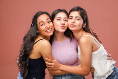 Sıradan kıyafetler giyen üç kadın birbirlerine sarılırken kameranın önünde somurtuyorlar.