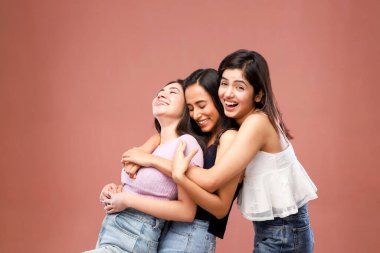 Üç güzel arkadaştan oluşan bir grup pembe bir arka planda dururken gülümsüyor ve birbirlerine sarılıyorlar.