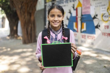 Sırıtkan, okul üniformalı, siyah bir sayfa tutan, omzunda bir çantayla kırsal bir bayan öğrenci.