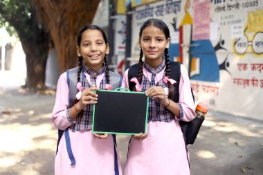 Gülümseyen Utangaç Kızlar ellerinde siyah bir tahtayla okulun arka planında duruyorlar.