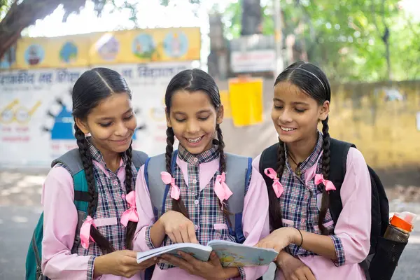 Gazeteyi verdikten sonra gelen üç Hintli bayan arkadaş okulun önünde duruyorlar ve biri elinde bir kitap tutuyor ve üçü de o kitaba bakıyor.