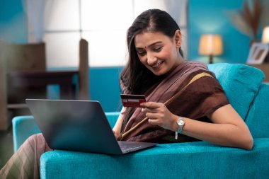 Hoş, olgun bir bayan dizüstü bilgisayarla kanepede oturuyor ve evde online alışveriş yaparken kredi kartı kullanıyor.