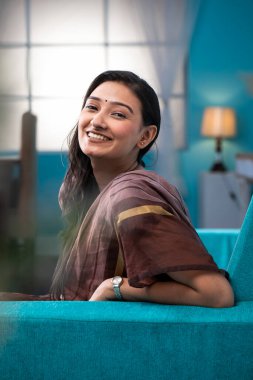 Mavi koltukta oturan, kameranın önünde poz veren, gülümseyen kadın.