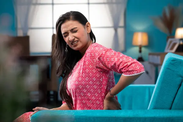 Olgun bir kadın koltukta oturuyor ve sırt ağrısı çekiyor. Sırtını eliyle tutuyor. 