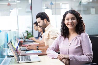 Kadınlar kameraya poz verirken, insanlar ofiste oturup projeleri üzerinde çalışıyorlar.