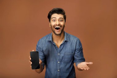 Mavi gömlekli genç bir adam kameraya doğru büyük bir gülümsemeyle akıllı telefon ekranını gösteriyor.