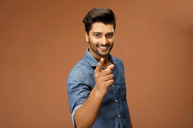 Mavi kot gömlekli neşeli bir adam parmağını kameraya doğru gösteriyor.