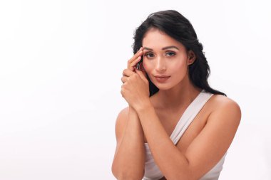 Siyah dalgalı saçlı zarif bir kadın beyaz tek omuzlu bir elbise içinde zarifçe poz verir..