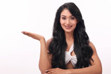 Gülümseyen bir kadın, uzun, dalgalı siyah saçlar ve parlak bir gülümsemeyle temiz beyaz bir arkaplanda poz verir. Bir elini nazikçe kaldırarak metin ya da reklam için mükemmel bir kopya alanı yaratıyor..