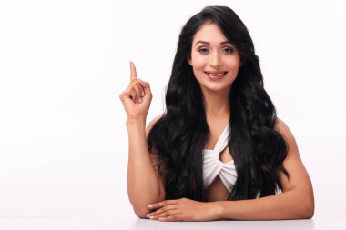 Uzun, dalgalı siyah saçları ve parlak bir gülümsemesi olan tatlı bir kadın beyaz bir arka planda oturuyor. Bir fikir sunar ya da bir noktayı vurgular gibi işaret parmağını kaldırır..
