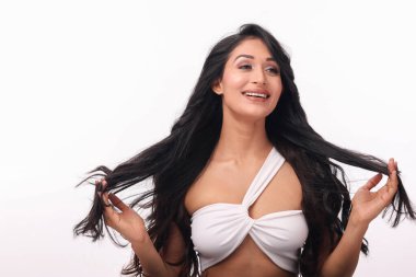 Uzun, dalgalı siyah saçlı canlı bir kadın saçlarını tutarken neşe içinde poz veriyor. Şık beyaz bir bluz giyip, temiz beyaz bir arka plana karşı özgüven ve cazibe yayıyor. Güzellik, moda ya da saç bakım reklamları için mükemmel..