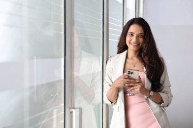 Bir kadın gülümseyen ve akıllı telefonunu parlak bir alanda kullanırken cam bir pencerenin yanında duruyor..