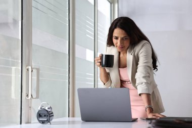 Spor ceketli bir kadın, elinde kahve fincanı olan bir bilgisayarın üzerine eğiliyor..