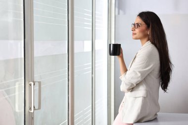 Bir kadın elinde kahve fincanıyla pencerenin yanında duruyor ve dikkatlice uzaklığa bakıyor..