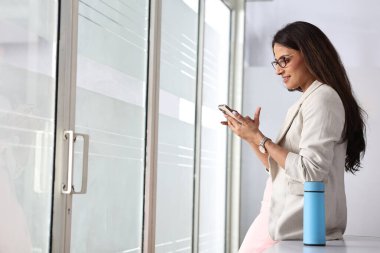 Bir kadın, parlak ve modern bir mekanda pencereye yaslanırken akıllı telefonunu kontrol eder..