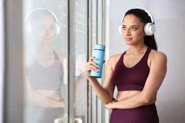 Genç bir kadın, elinde su şişesiyle pencereden dışarı bakarken kulaklıkla müzik dinlemekten hoşlanır..