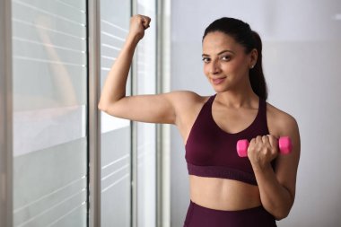 Kendine güvenen bir kadın, parlak bir spor salonunda pembe bir halteri tutarken kaslarını esnetir..