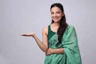 Bir kadın yeşil sari giyerken gülümser ve elini davetkar bir şekilde uzatır..