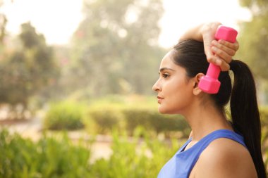 Parkta dikilip dambıl egzersizi yapan, mavi kısa bluzlu, formalı bir kız..