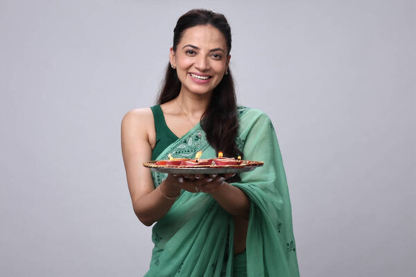 Женщина улыбается, держа в руках декоративную тарелку со сладостями и фруктами, одетую в зеленое сари.