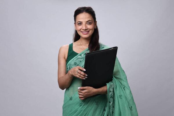 Женщина в зеленом сари уверенно стоит, держа папку, улыбаясь зрителю.