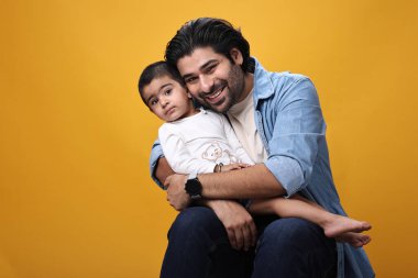 Bir baba, parlak sarı bir arka plana karşı çocuğunu sıcak bir kucaklama içinde gülümser..