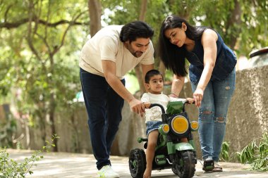 Bir çift, yeşilliklerle çevrili yolda üç tekerlekli bisiklete binen çocuklarına yardım ediyor..