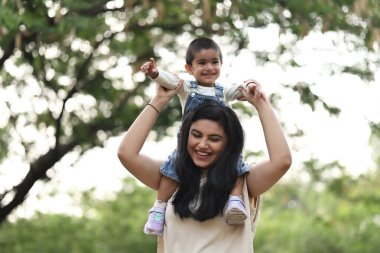 Neşeli bir anne, yeşillik dolu güneşli bir parkta küçük çocuğunu omuzlarında taşır..