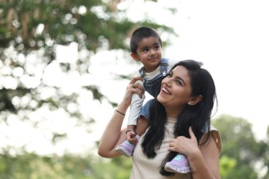 Anne, küçük oğlunu omuzlarında taşıyor. İkisi de ağaçlarla çevrili güneşli bir parkta gülümsüyor..