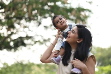 Neşeli bir anne, gülümseyen çocuğunu güneşli bir öğleden sonra yeşil bir parkta omuzlarında taşır..