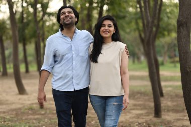 İki insan, akşam boyunca huzurlu bir parkta yeşil ağaçlarla çevrili bir şekilde gülümseyerek yürür..