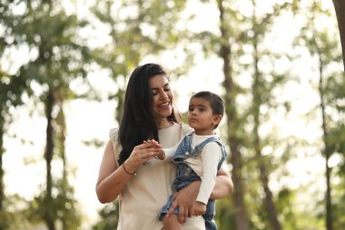 Bir anne, yeşil ağaçlarla çevrili güneşli bir parkta gülümserken oğlunu kucağına alır..