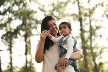 Sıcak ve güneşli bir günde ağaçların altında gülümseyen bir kadın oğlunu kucağına alır..