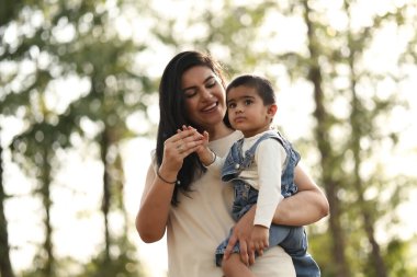 Güneşli bir günde yemyeşil bir ortamda gülümseyen bir anne çocuğunu kucağına alır..